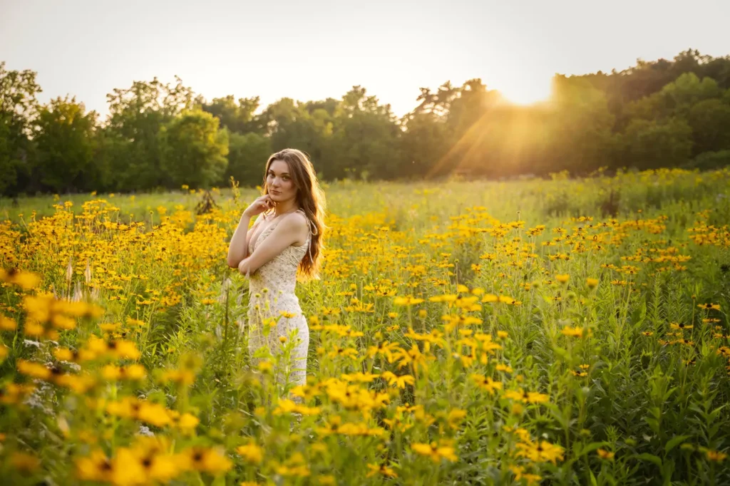 High school senior girl standing in a wildflower field at sunset at Aldo Leopold Nature Center, wearing a floral dress and softly smiling toward the camera.